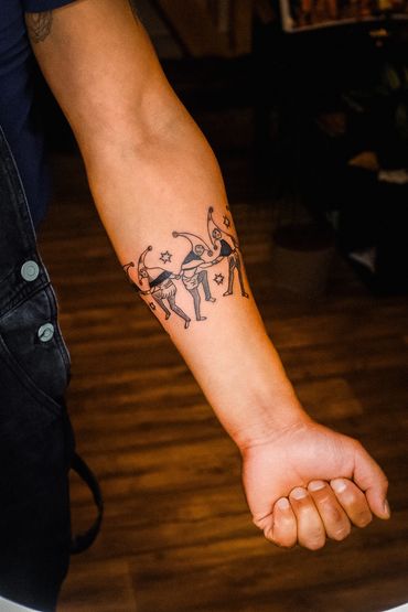 Tattoo of dancing jester figures on a forearm with stars around them.
