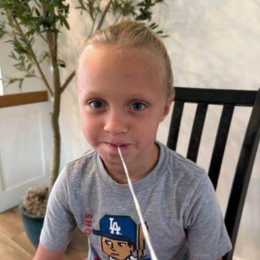 Young child with light hair playing with a string in their mouth indoors.