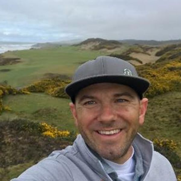 Smiling man wearing a gray cap outdoors on a cloudy day.