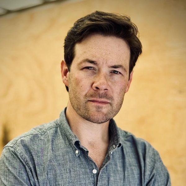 Man with light stubble wearing a blue shirt looks thoughtfully at the camera.