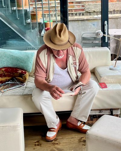 A man in a hat reads a book while sitting on a modern white couch indoors.