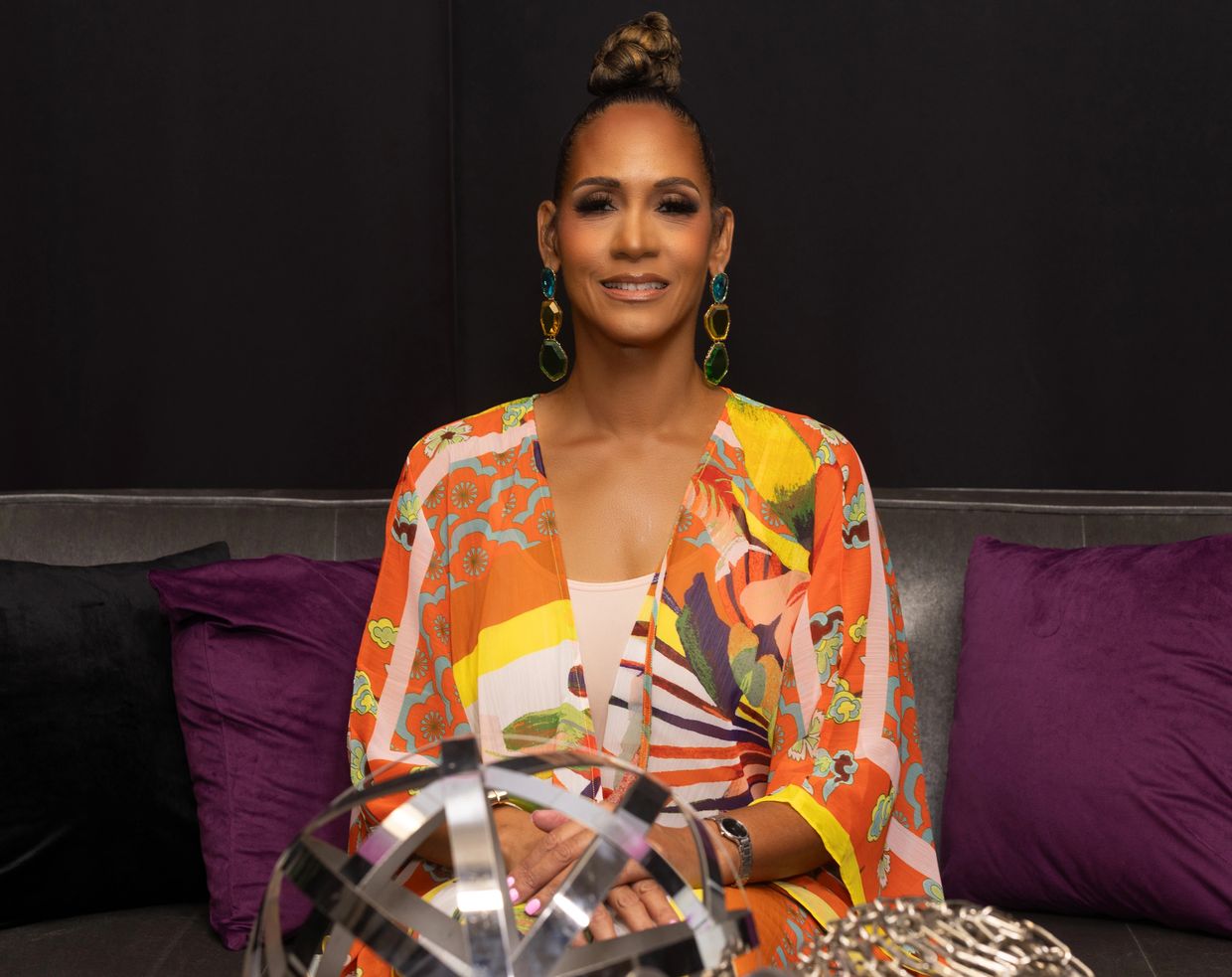 Woman in vibrant outfit sitting on a couch with purple pillows.