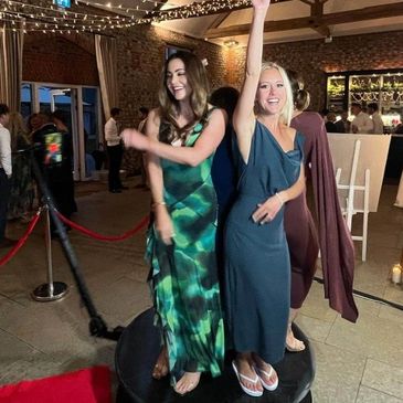 Two women in elegant dresses dancing on a rotating platform at a festive indoor event.