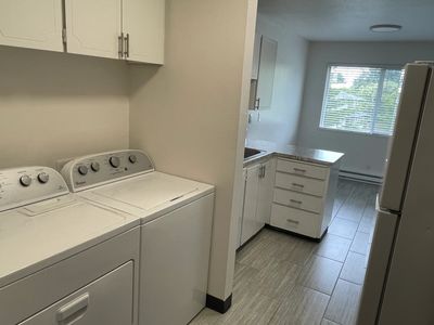 A laundry area with a washer and dryer next to a kitchen with white cabinets and a window.