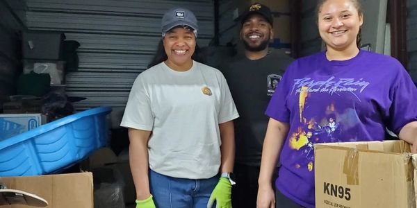 Three people smiling in a warehouse with boxes and gloves.