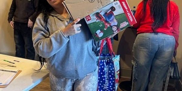 Two women, one holding a boxed toy and bags at a table indoors.