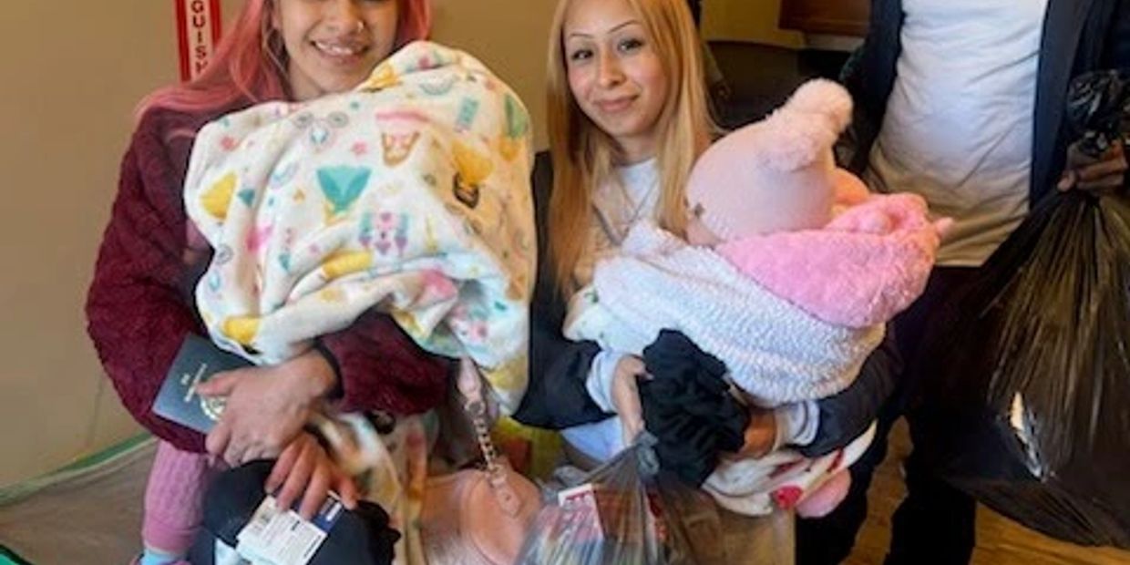 Two women each holding a baby wrapped in blankets, smiling indoors.