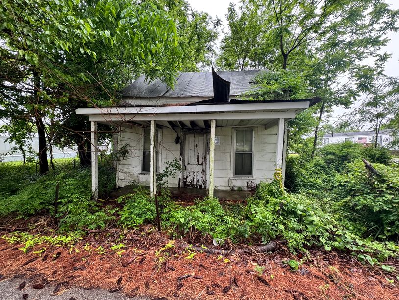 Abandoned, overgrown house with peeling white paint and a damaged roof.