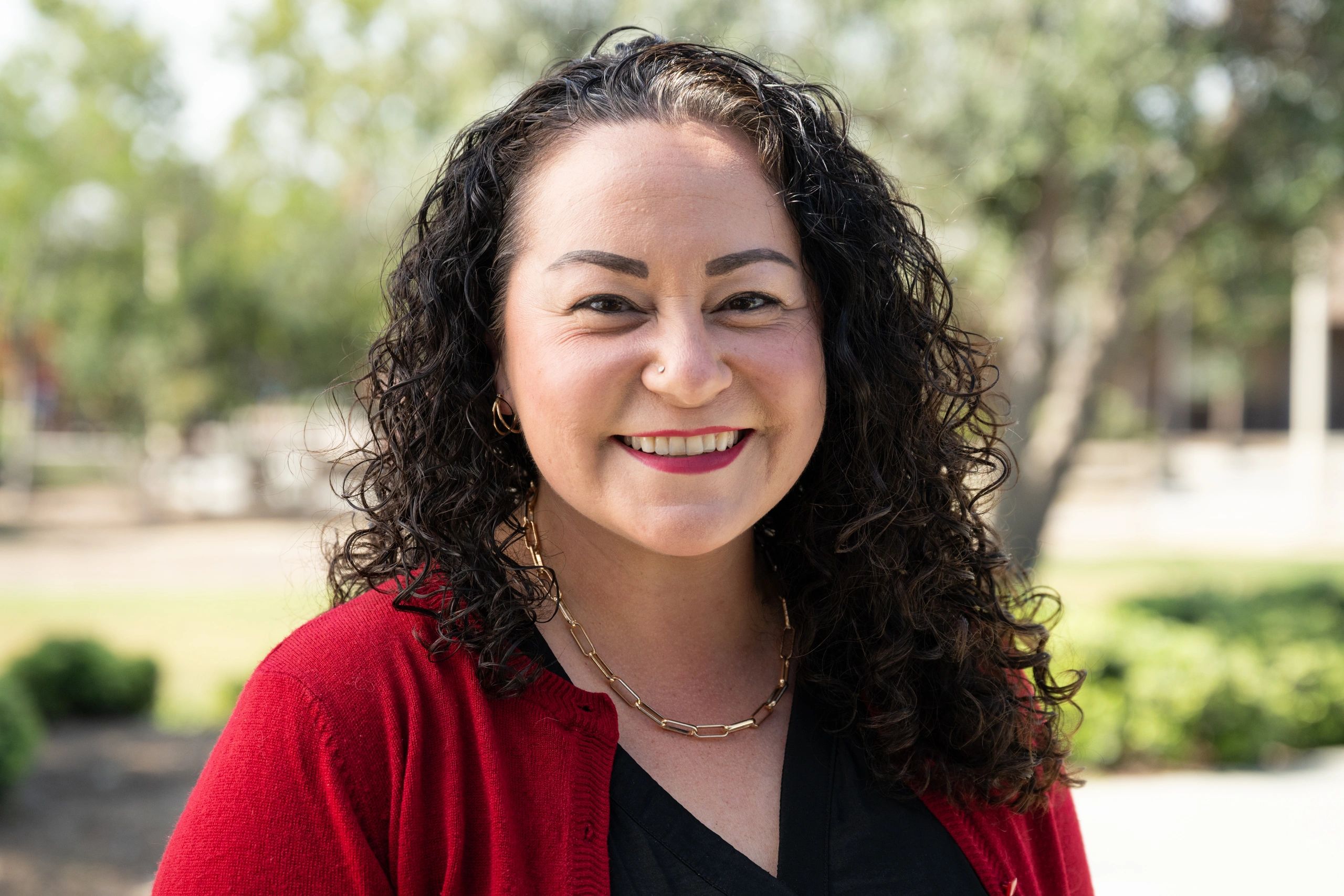 Smiling woman with curly hair wearing a red cardigan outdoors.