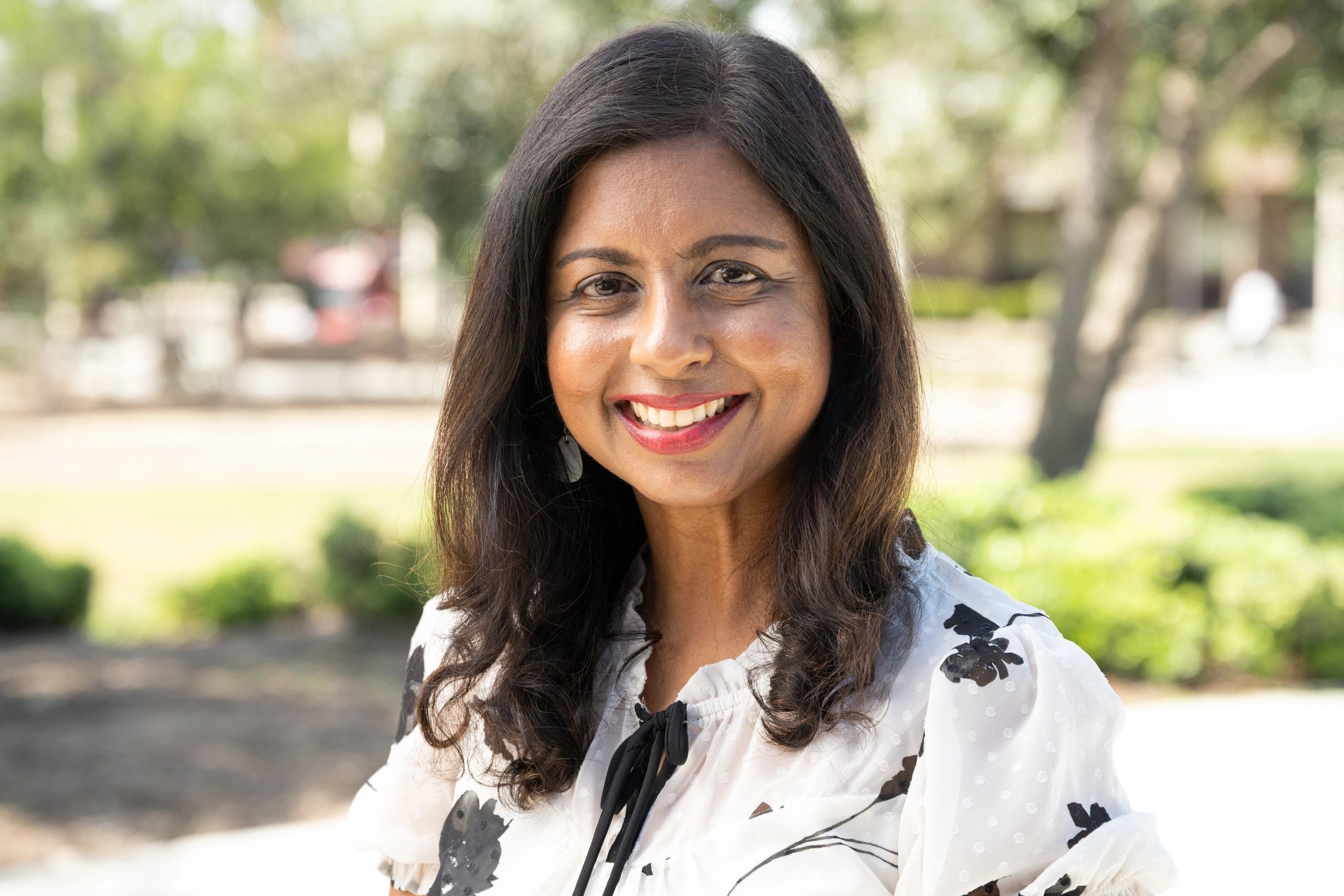 Smiling woman with long dark hair in a white floral blouse outdoors.