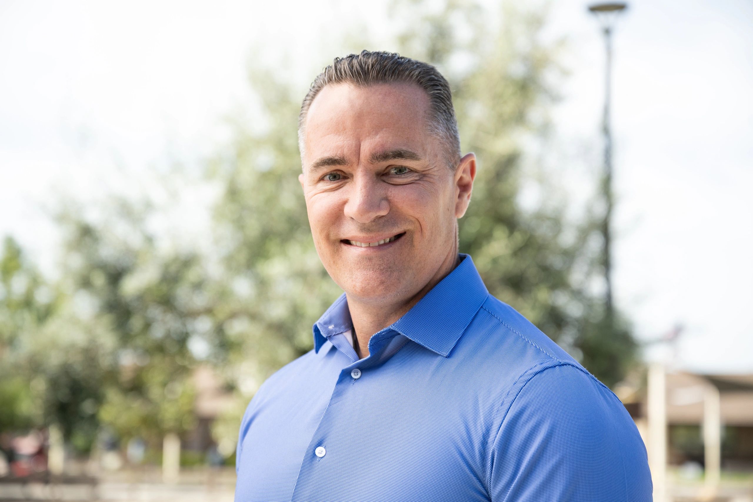 Smiling man in a blue shirt outdoors with a blurred natural background.