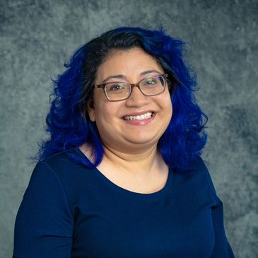 Smiling woman with blue hair and glasses against a textured backdrop.