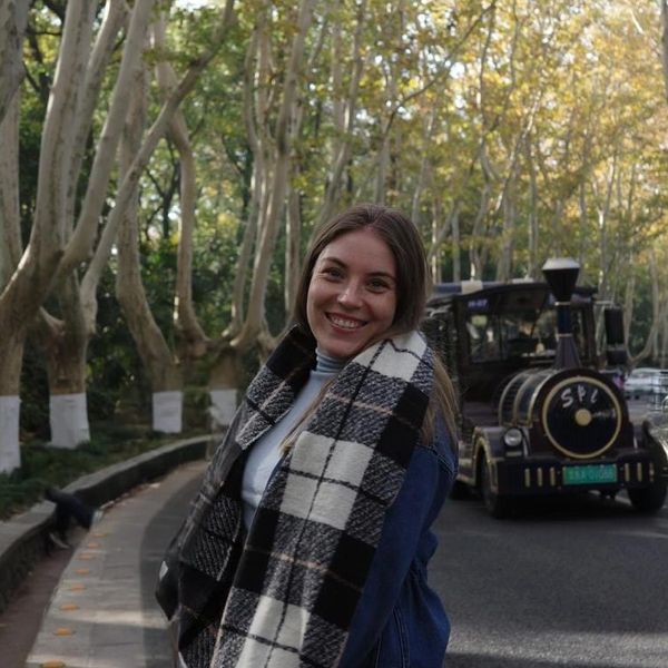 A smiling woman with a plaid scarf stands on a tree-lined road with a vintage train behind her.