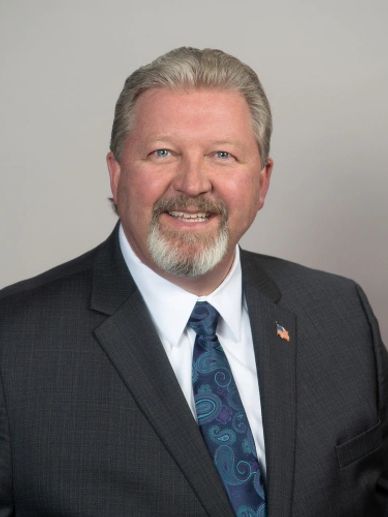 Smiling man in a dark suit with a patterned blue tie and small flag pin.
