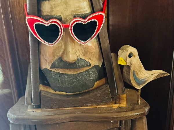Wooden face sculpture with heart-shaped sunglasses and a small bird figurine.