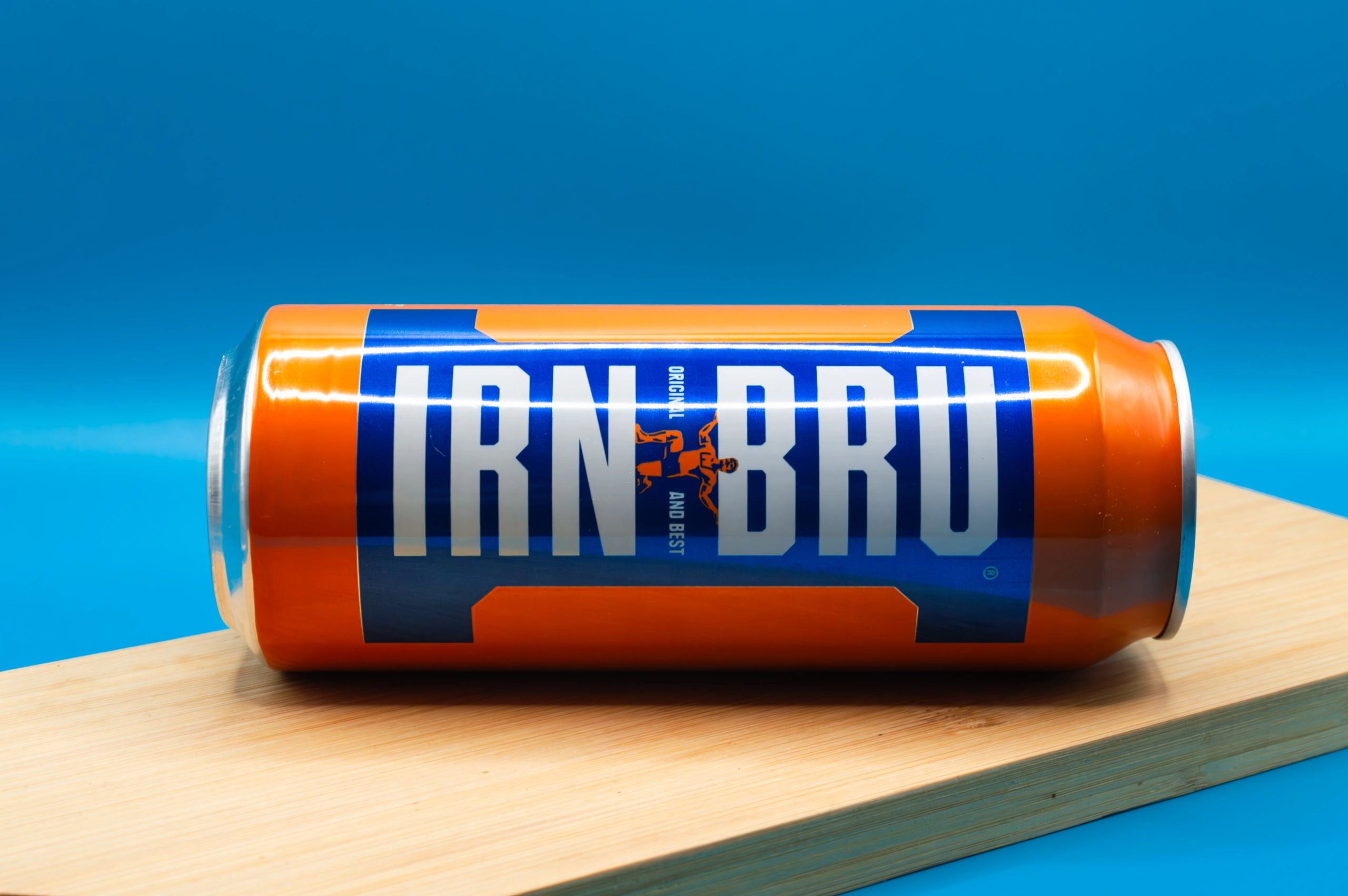 Orange and blue Iron Bru can on a wooden surface against a blue background.