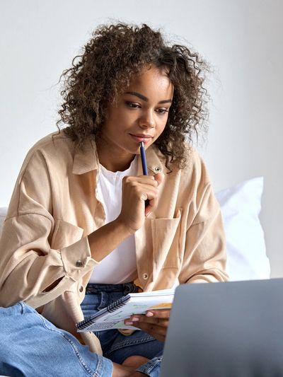 A young woman thoughtfully working on a laptop with a notebook and pen.