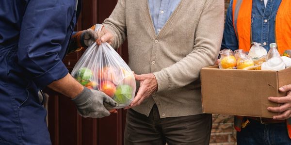 Two workers deliver groceries to a happy elderly man at his home.