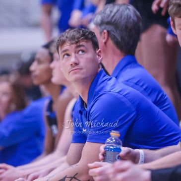 A young man in a blue shirt looks up while sitting among a group of people in a sports setting.