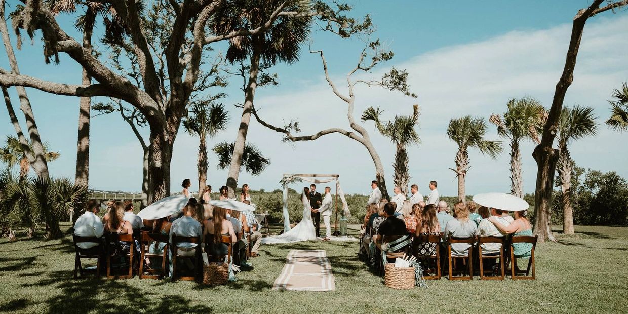 Outdoor wedding ceremony under large trees with guests seated in rows.