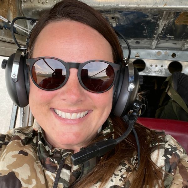 Smiling woman with sunglasses and headset inside an aircraft cockpit.