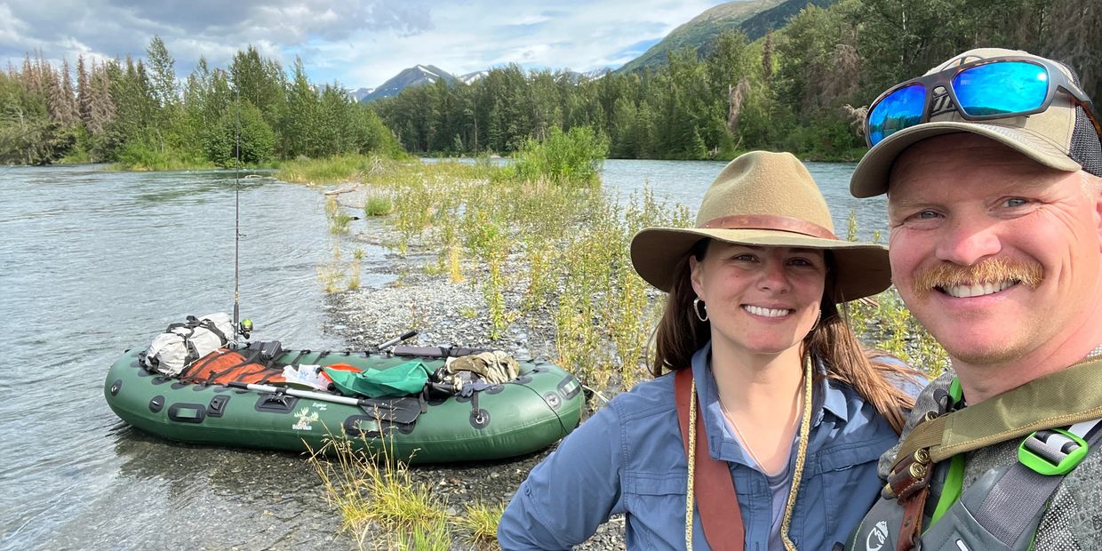 A smiling couple stands by a river with a packed inflatable raft in the background.