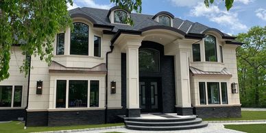 Beige two story big house with black trim and lawn and trees on a sunny day