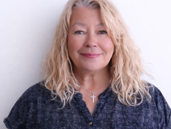 Smiling middle-aged woman with blonde curly hair wearing a dark top and cross necklace.