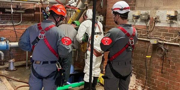 Workers in safety gear inspect an industrial pipe system in a confined space.