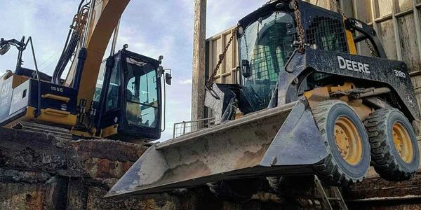 An excavator lifting a skid steer out of a construction pit in a manufacturing facility. 