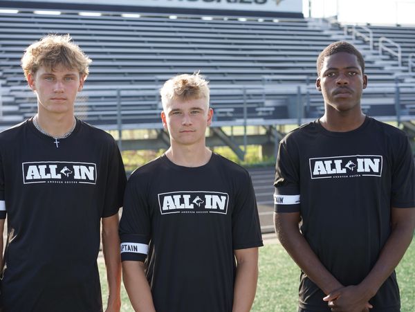 Three young male soccer players standing on a field wearing black "ALL IN" shirts.