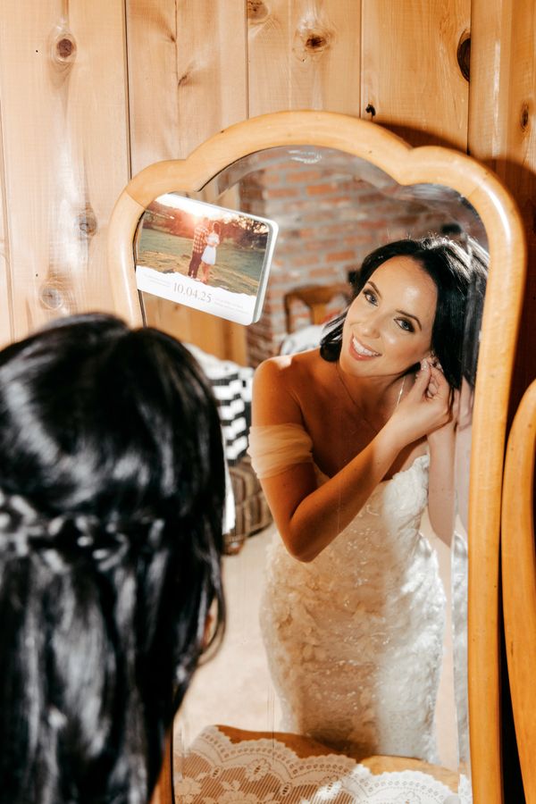 A bride adjusts her earring while looking into a wooden mirror.