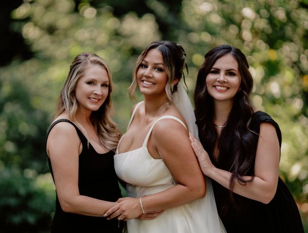 Three women smiling, bride in white gown, friends in black dresses.