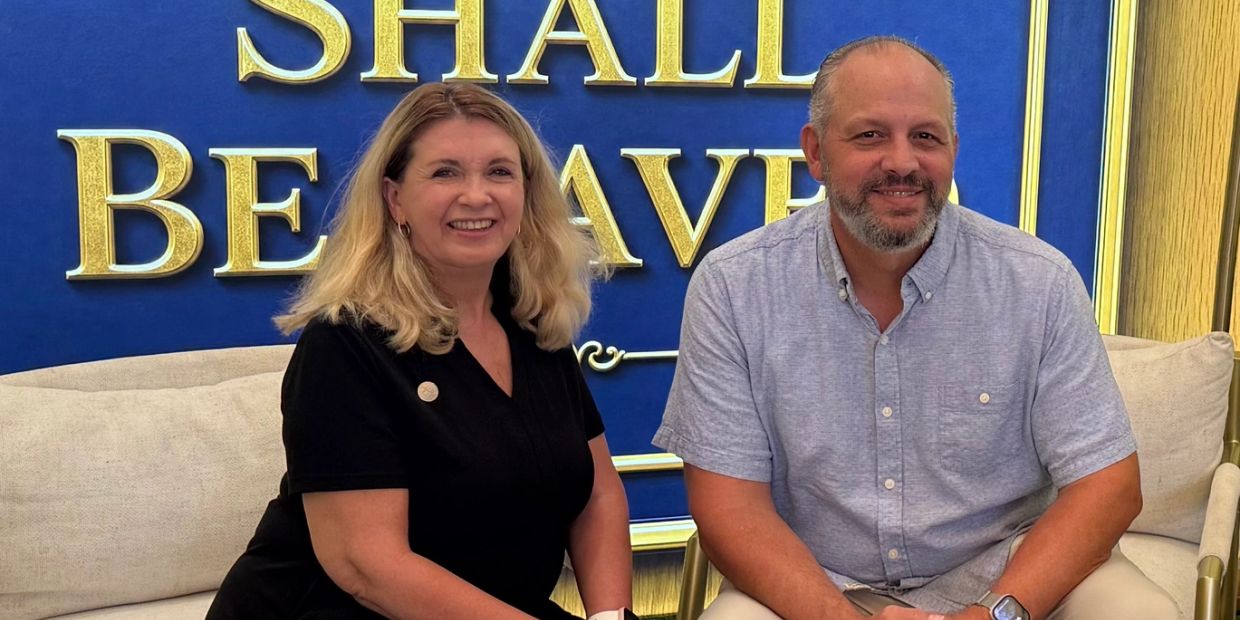 Two people sitting and smiling in front of a blue and gold sign.