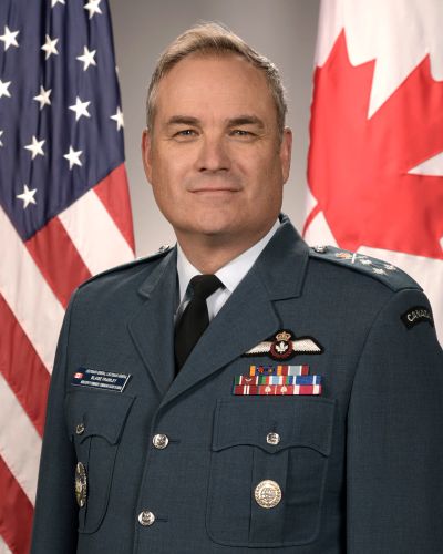 Military officer in uniform with US and Canadian flags behind.