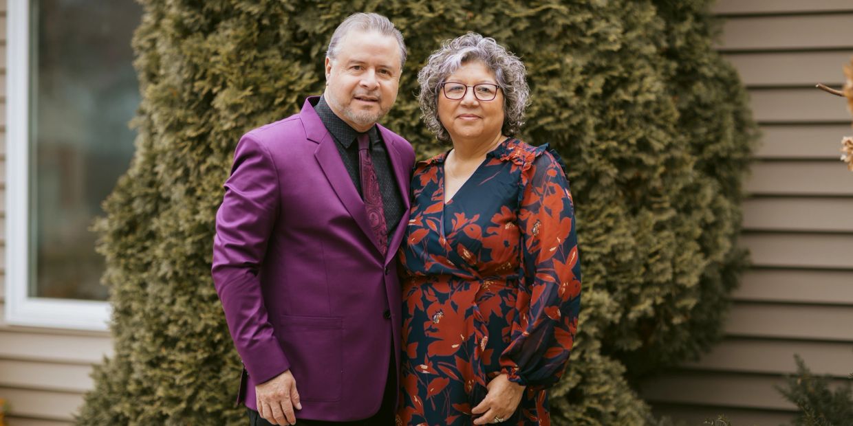 A well-dressed couple posing outdoors in front of greenery.