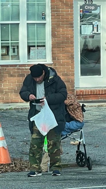 Man in a black jacket and camo pants standing outdoors with a plastic bag.