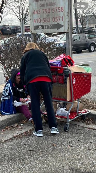 A person helping a homeless individual on a sidewalk with a shopping cart nearby.