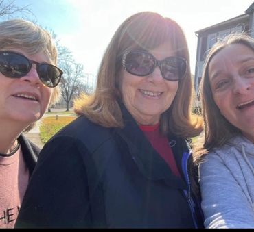 Three women wearing sunglasses smiling outdoors on a sunny day.