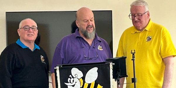 Three men standing and singing around a microphone with a bee-themed banner.
