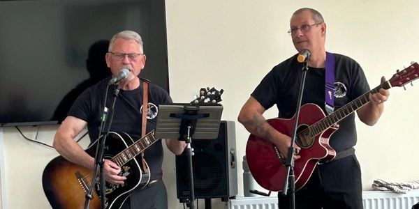 Two men playing acoustic guitars and singing into microphones indoors.