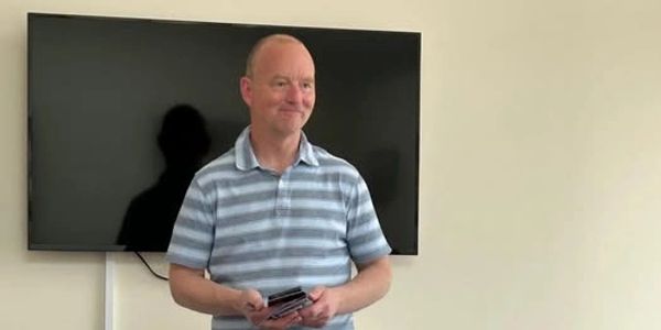 A man in a striped shirt stands in front of a large TV, smiling.