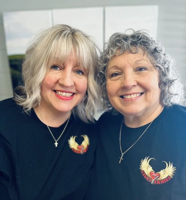 Two women smiling, wearing matching black shirts with angel wings and heart design.