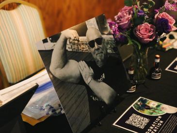 Black and white photo of a man flexing his arm on a table with flowers and donation info.