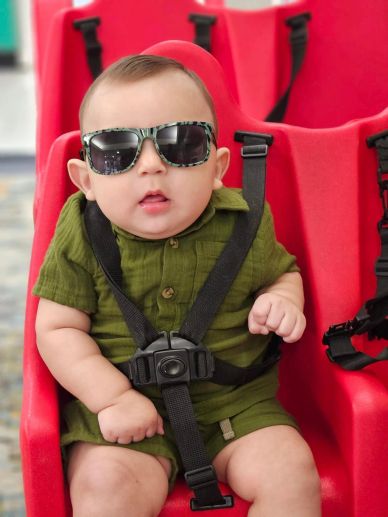 Baby wearing sunglasses and green outfit sitting in a red seat with harness.