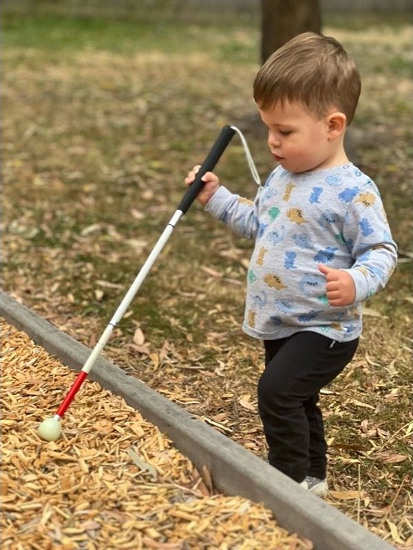 A young child exploring the step up to a playground with a long cane. 