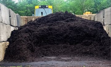 Large pile of dark mulch stored between concrete walls with machinery behind.
