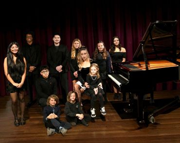 Group of young musicians posing by a grand piano on stage.