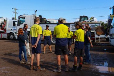 Workers in yellow shirts stand in a circle outdoors near trucks.