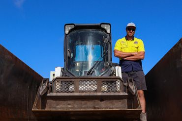 Man standing confidently beside a large construction vehicle under a clear blue sky.